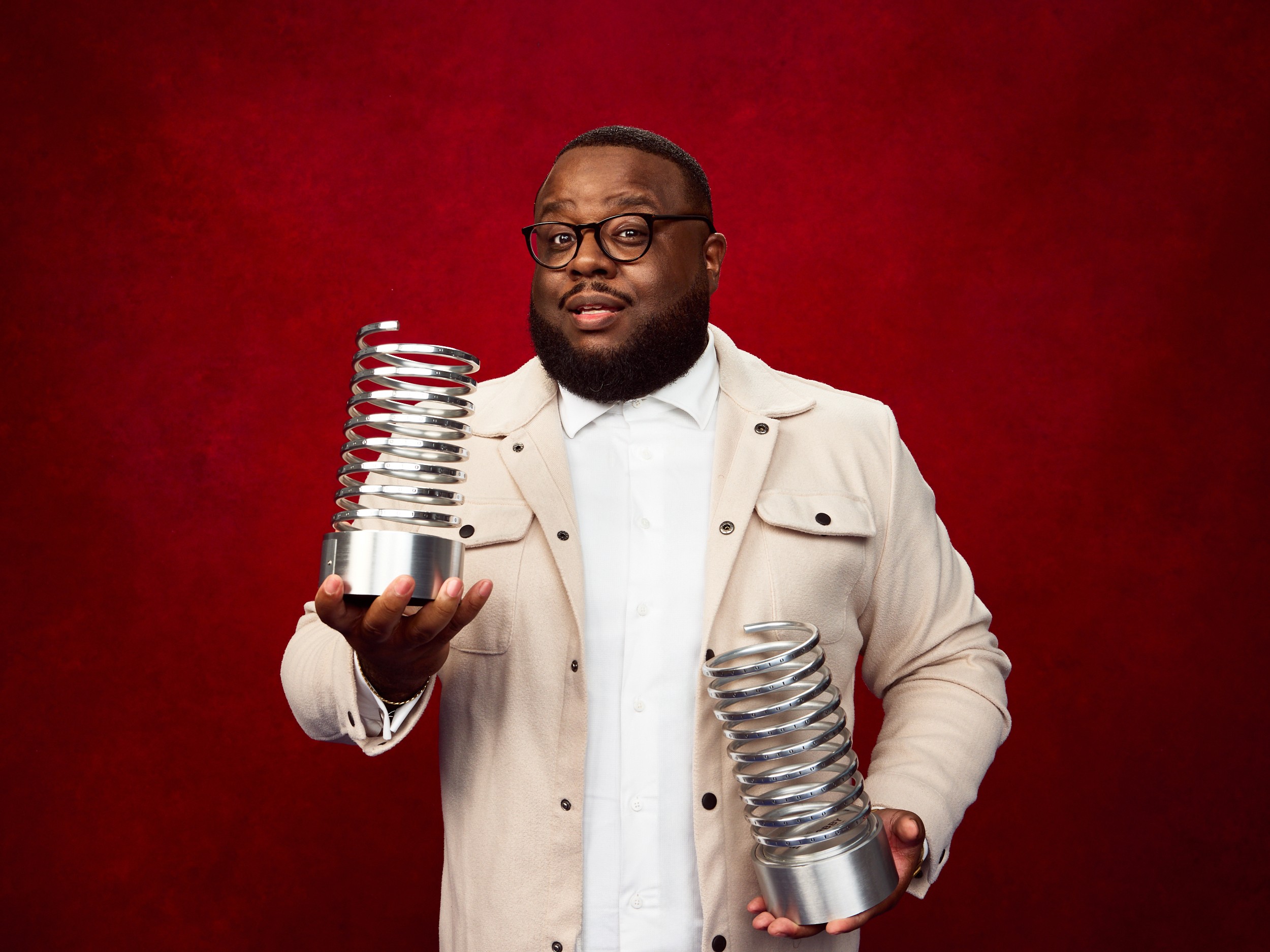 Man with glasses and beard holding metal coil objects against a red background.