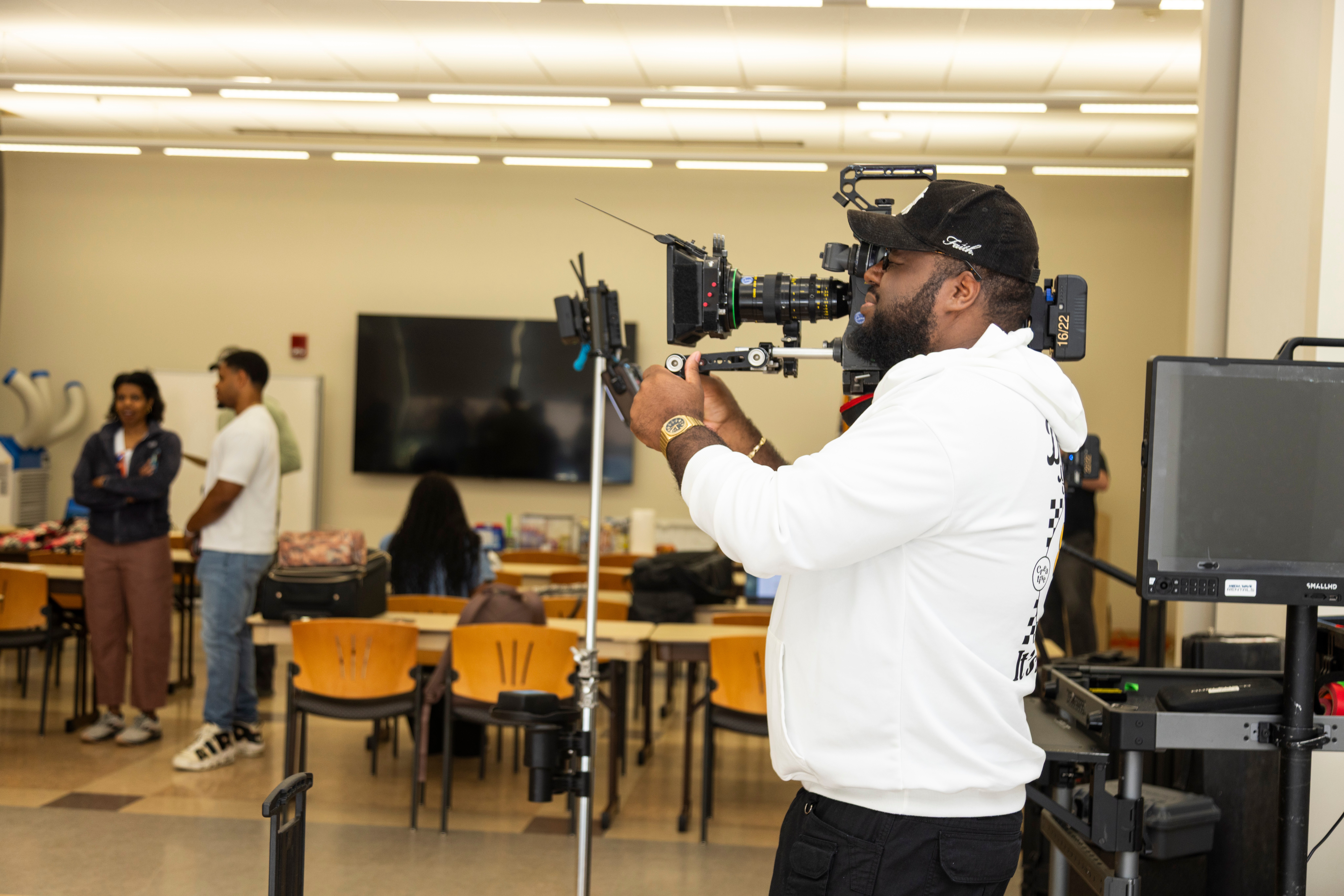 A man with a camera on his shoulder films a group of people in a classroom or conference room setting.