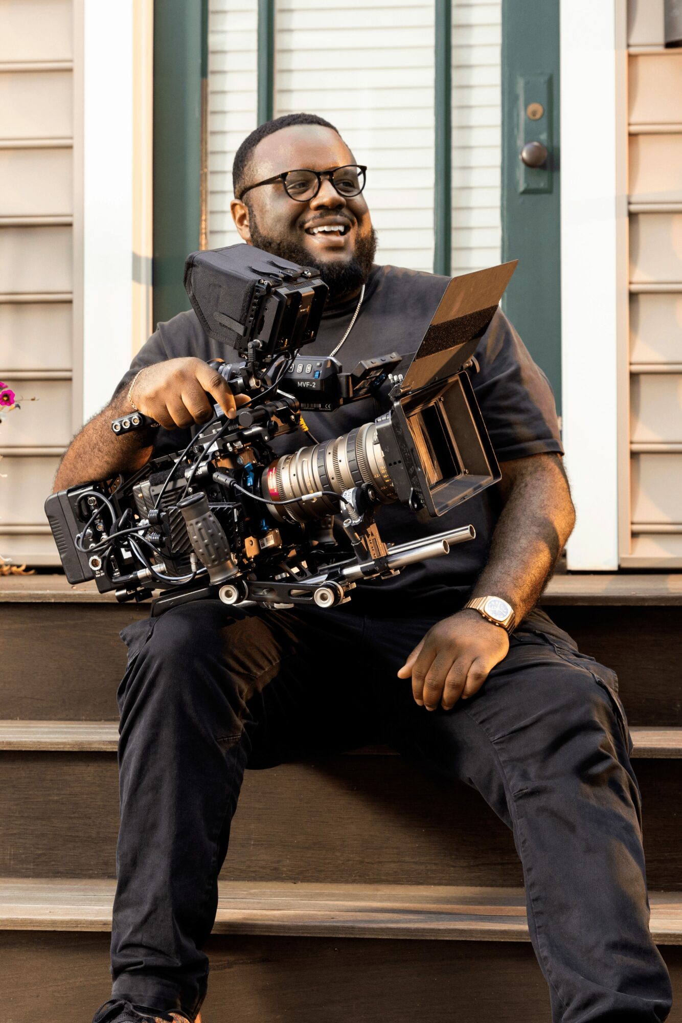 Man sitting on steps holding professional camera equipment, smiling, in front of a house with window shutters.