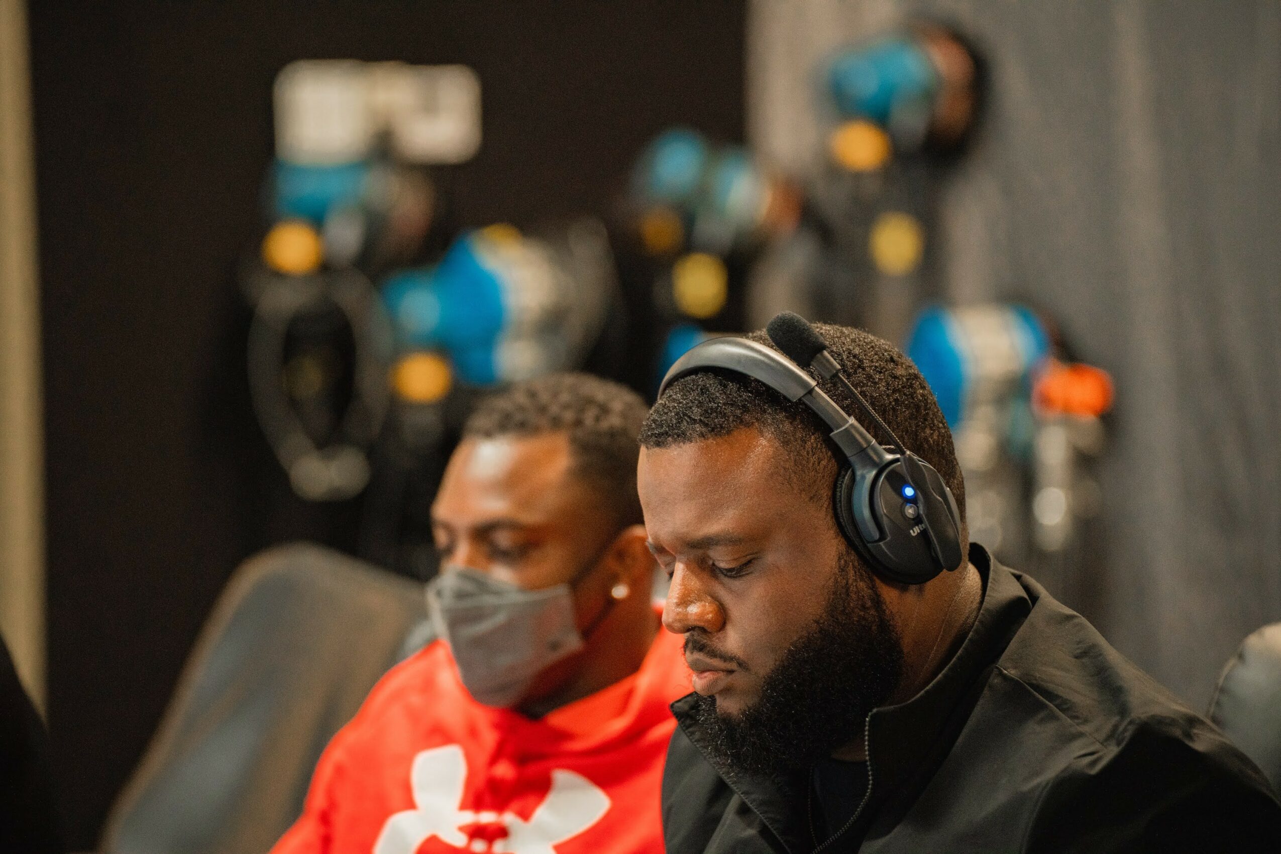 Two men wearing headphones and face masks sit in a dark room with equipment in the background.