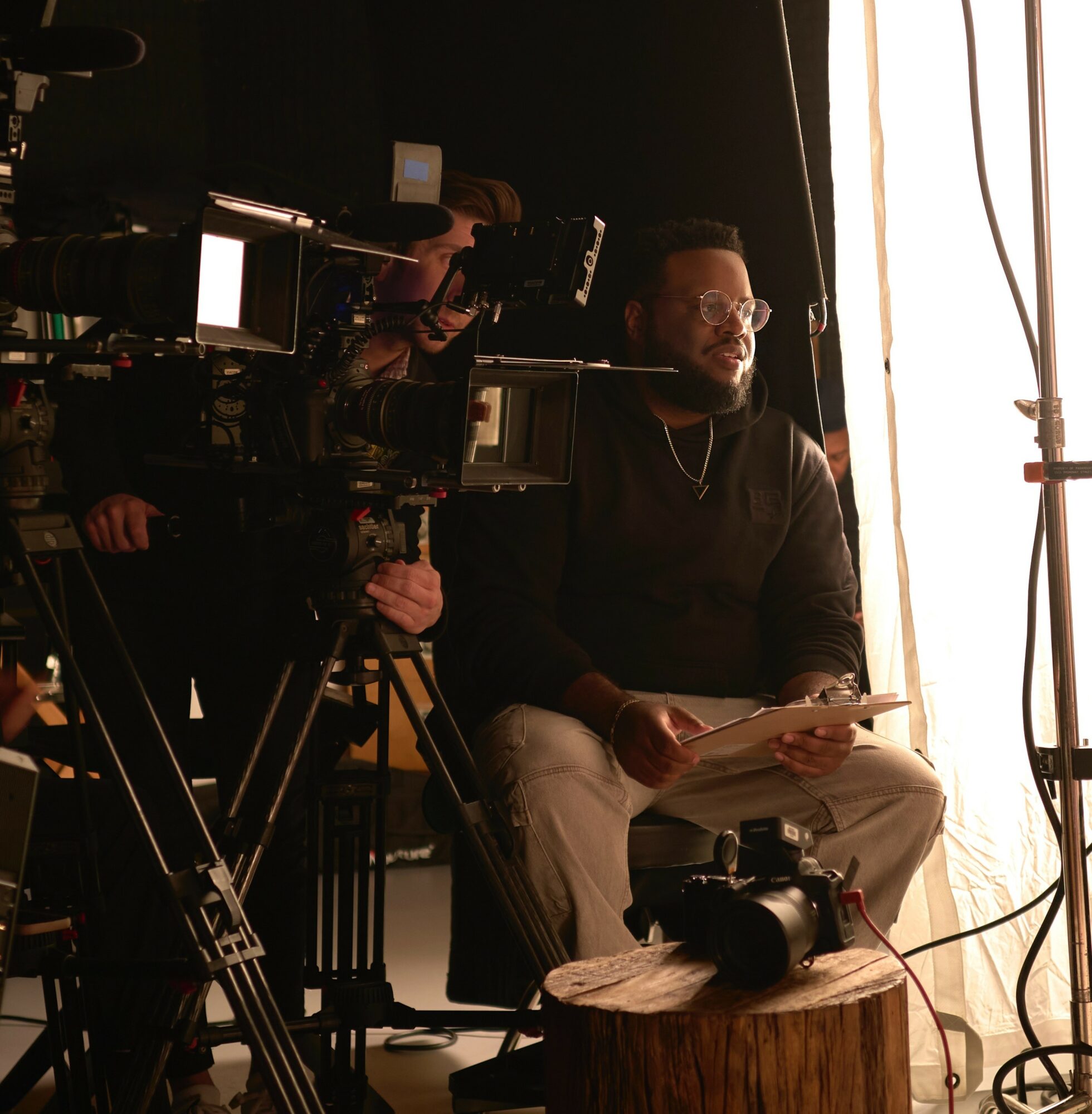 Man sitting with papers, filming equipment around him, in a studio with bright light from a window, dark background.