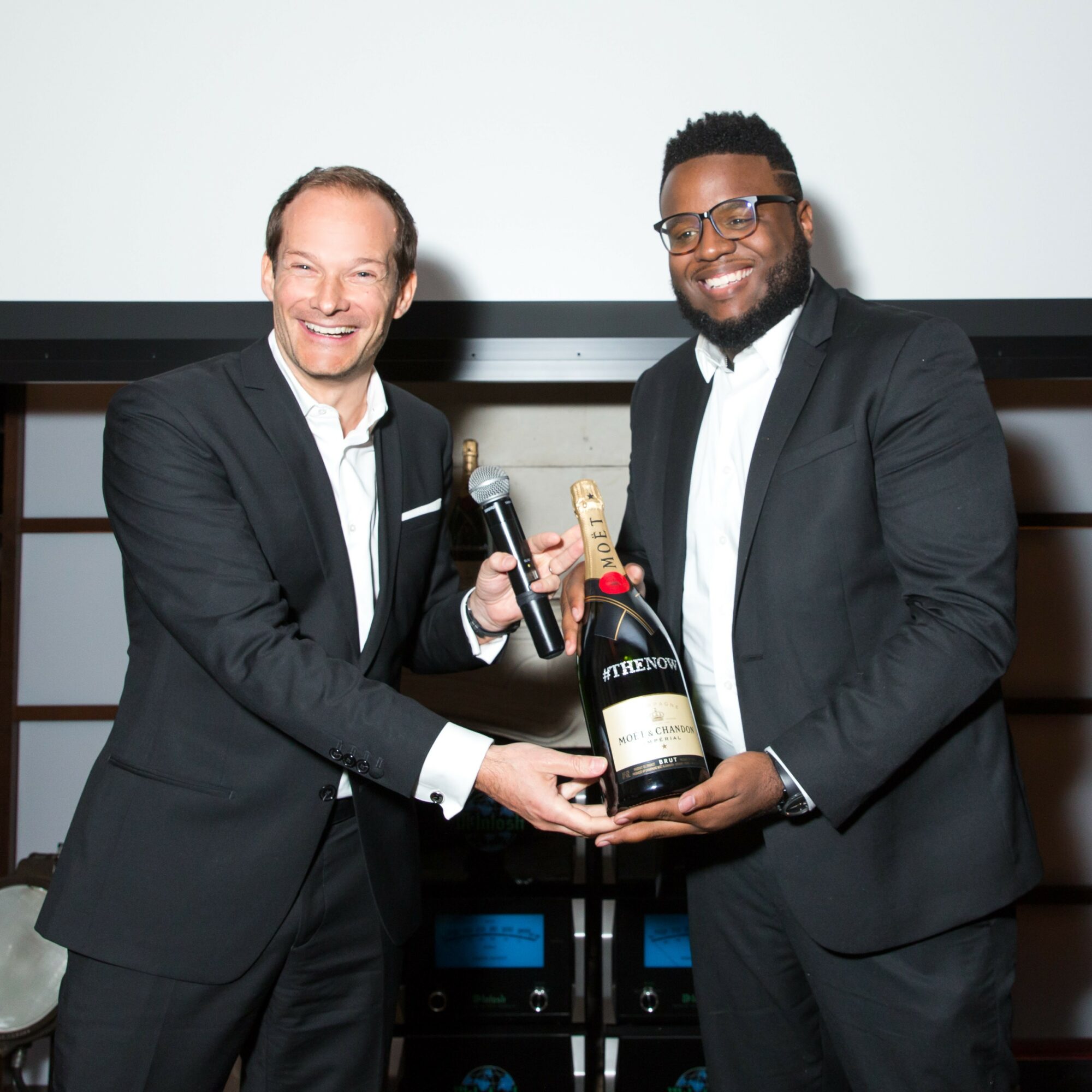 Two men in suits smiling, holding a large bottle of champagne and a microphone, standing in front of a white background.