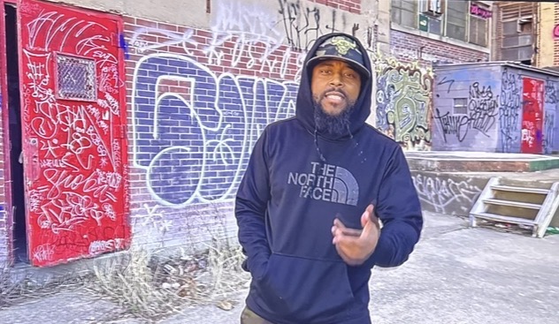 Man wearing a hoodie and helmet standing in front of graffiti-covered wall, holding a phone, outdoors in winter.