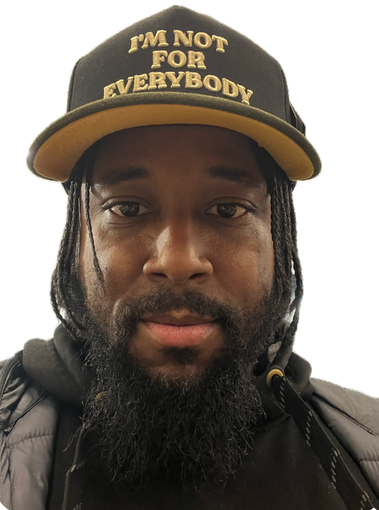 Close-up of a man wearing a black cap with gold text, black jacket, and dreadlocks, looking at the camera.