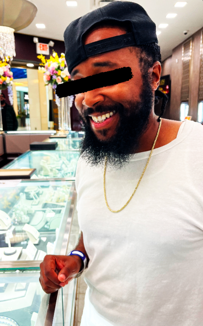 Smiling man with a beard, wearing a black cap backwards, white shirt, gold chain, in a jewelry store.