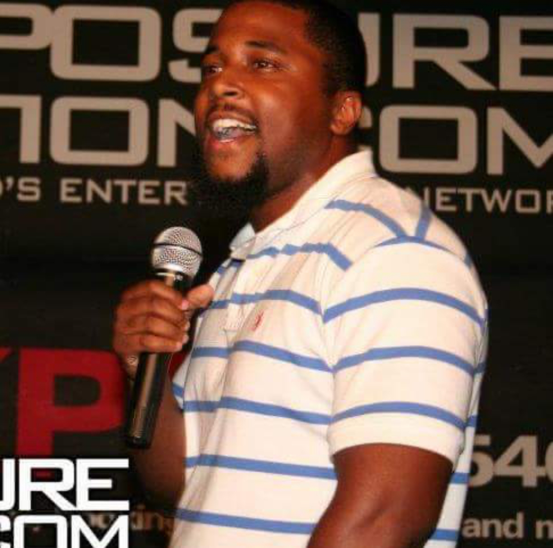 Man speaking into a microphone, wearing a white and blue striped shirt, smiling, with a black background and text.