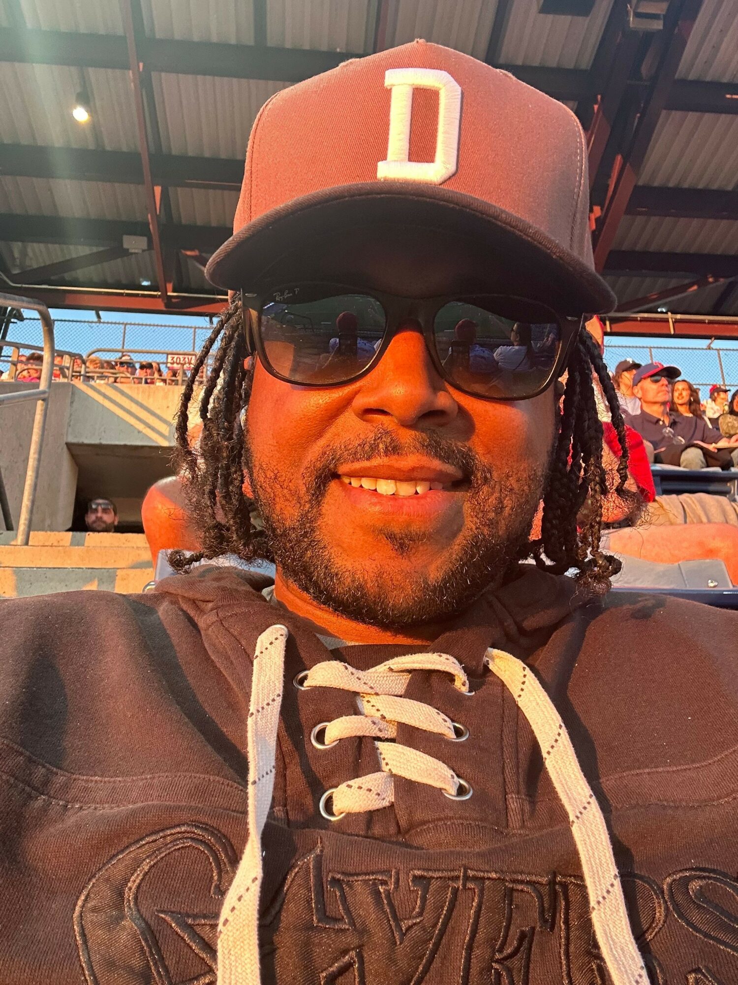 Person wearing a baseball cap, sunglasses, and hoodie, smiling outdoors with a cityscape background.