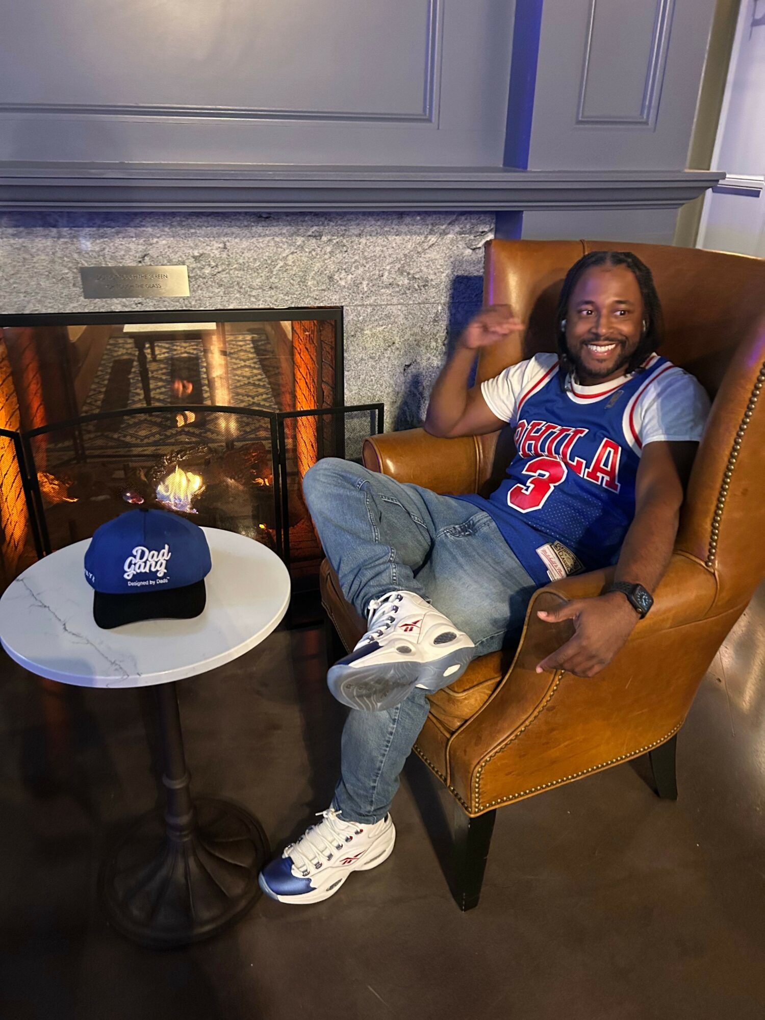 Person sitting in a leather chair, wearing a sports jersey, jeans, and sneakers, in a room with a fireplace and a small table.
