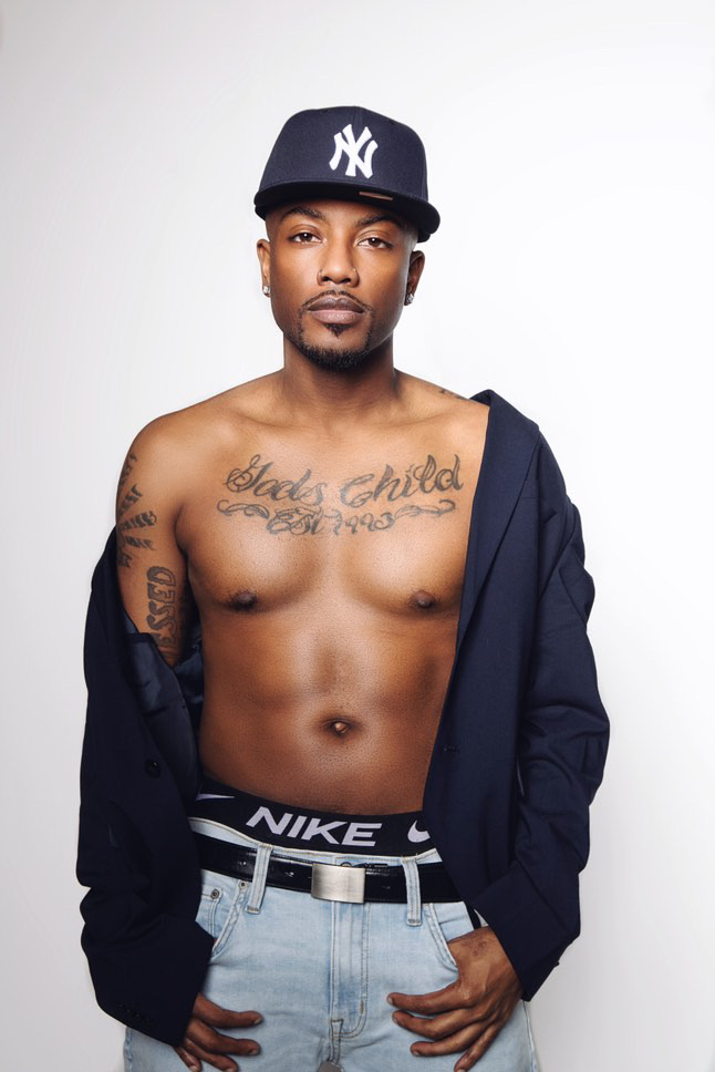 Young man with a cap, shirt partially off, showing tattoos on chest and arm, standing against a plain background.