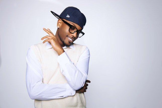 Young man wearing glasses and a baseball cap, smiling with eyes closed, arms crossed, against a plain background.