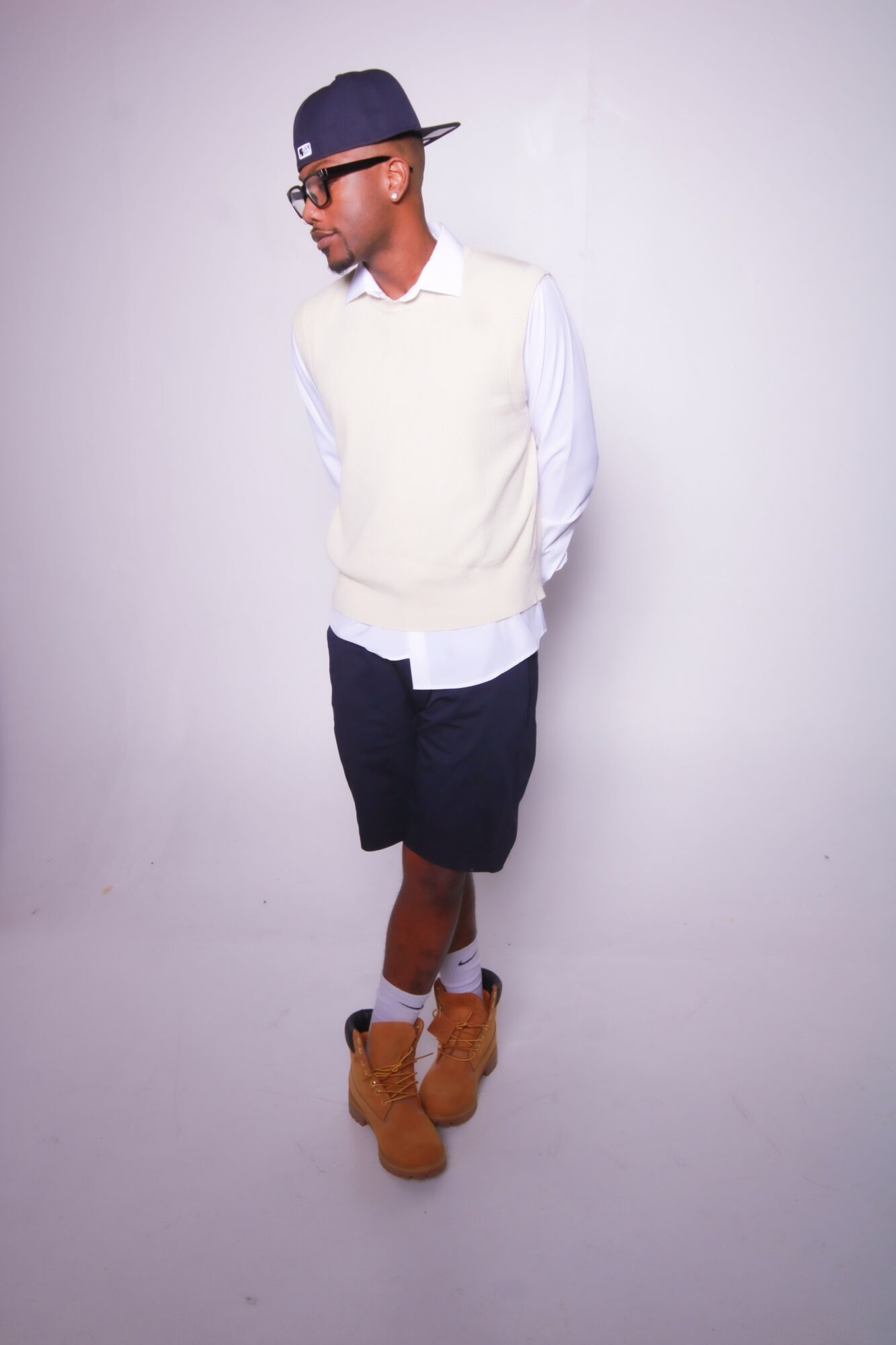 Young man standing with hands behind back, wearing glasses, a cap, white shirt, light sweater, dark shorts, and tan boots.