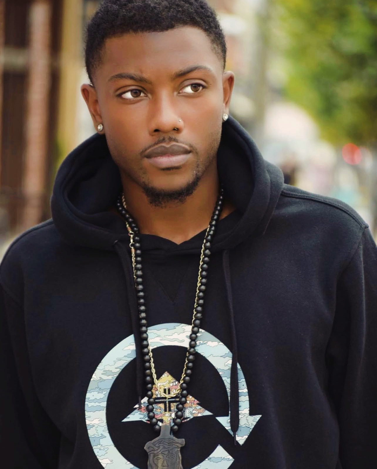 Young man with short hair and earrings wearing a black hoodie and necklace outdoors.