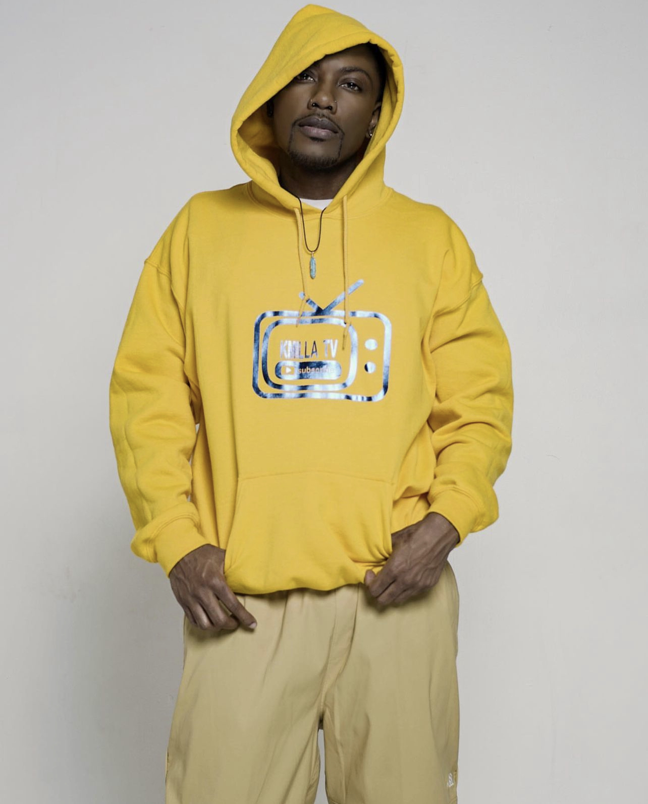 Man wearing a yellow hoodie with a graphic on the front, beige pants, standing against a plain background.