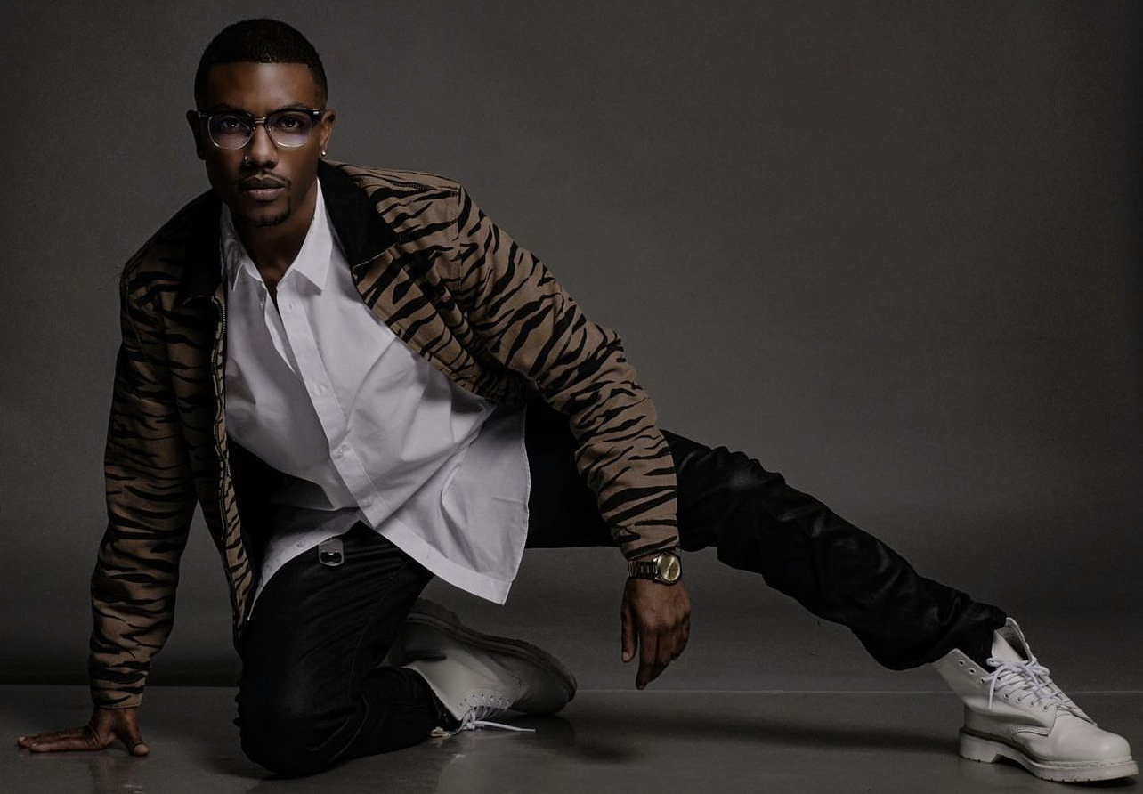 Person in white shirt and tiger-striped jacket crouching on the floor with one hand and leg extended, against a dark background.