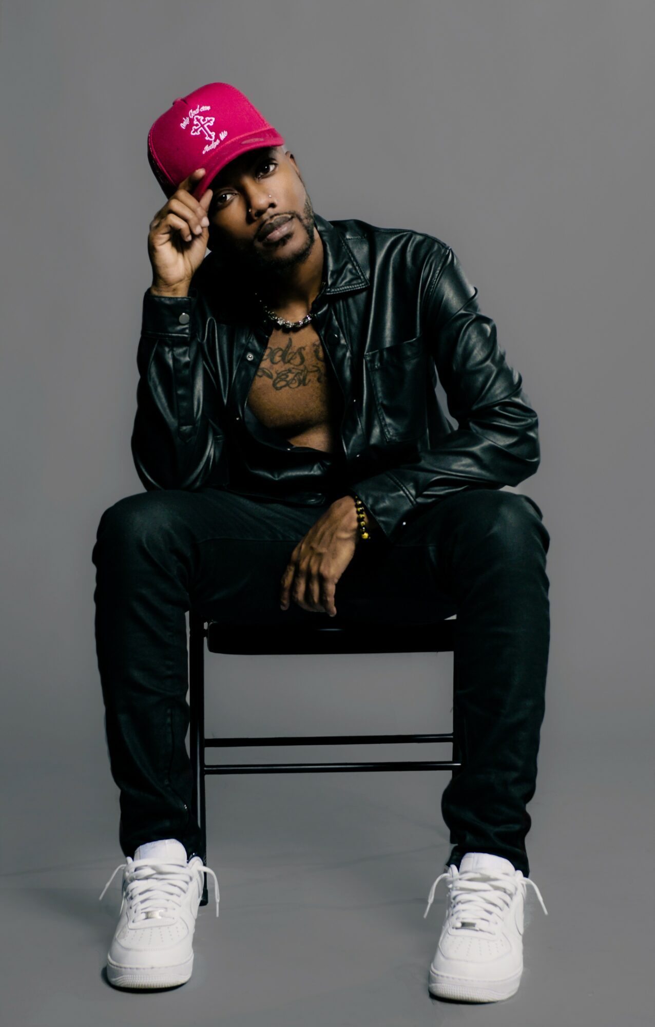 Man sitting on a chair wearing a black leather jacket, pink cap, white sneakers, and jewelry, against a gray background.