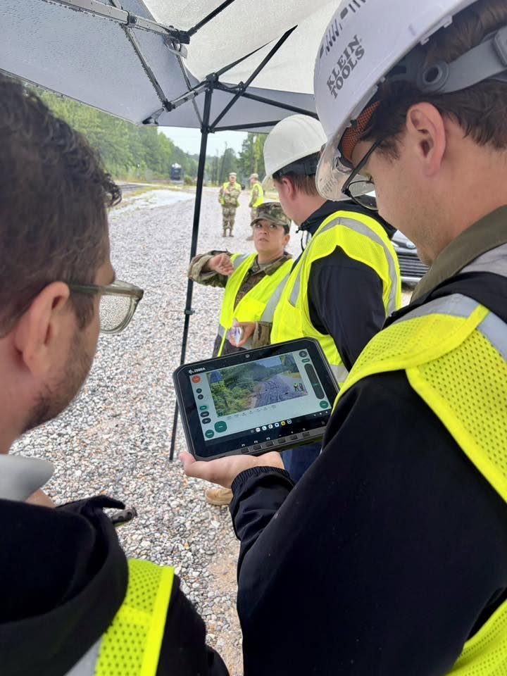 Group of people wearing safety vests and helmets outdoors, one person holding a tablet displaying a map, under an umbrella.