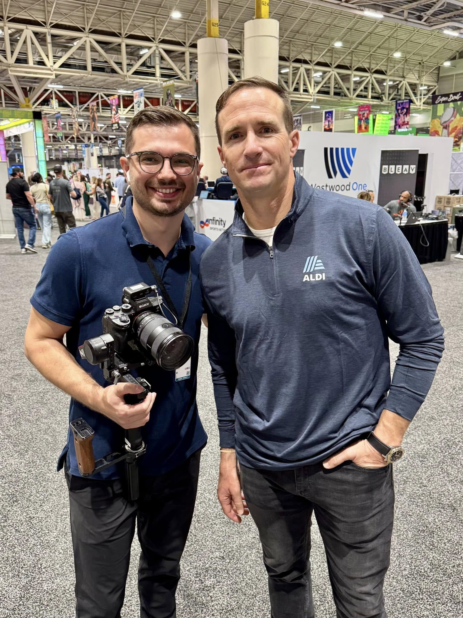 Two men standing together at an indoor event, one holding a camera, both smiling, with booths and people in the background.