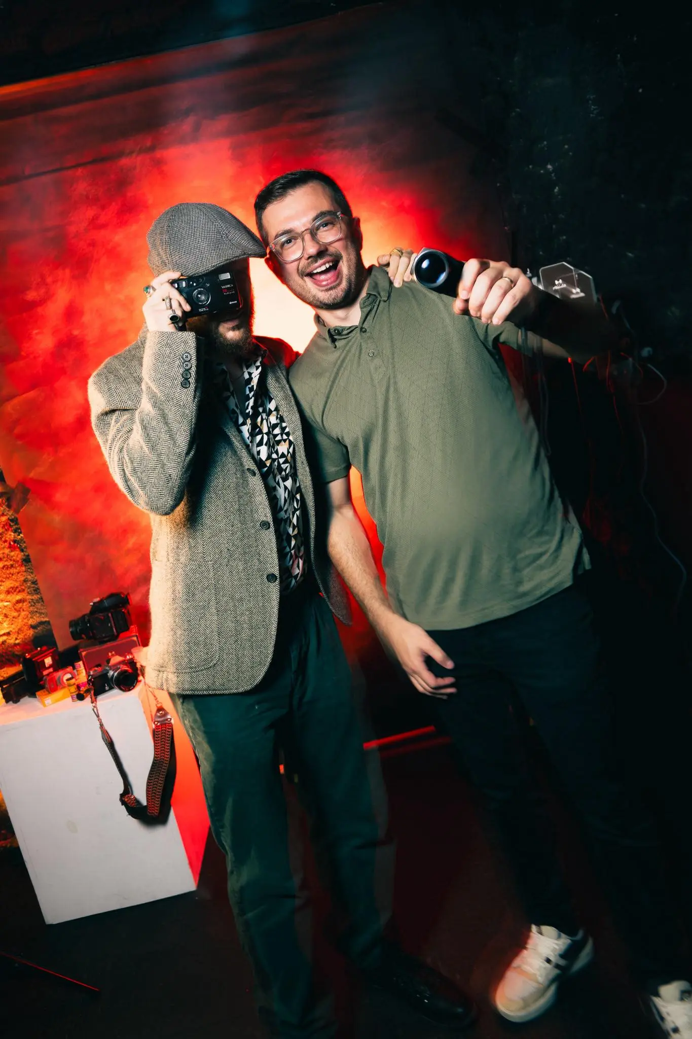 Two men smiling, one holding a camera, in a dimly lit room with red lighting background.