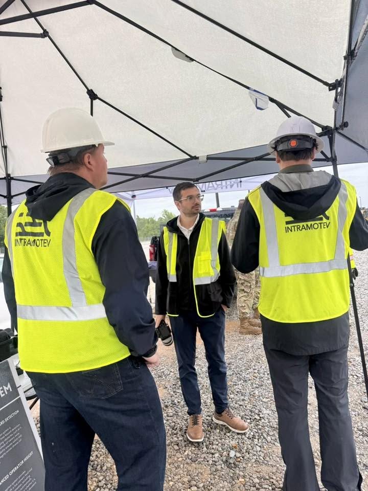 Three men in yellow safety vests and hard hats under a tent, engaged in conversation outdoors.