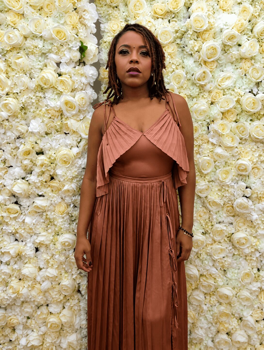 Woman in brown dress standing in front of a white floral wall, looking at the camera with a serious expression.