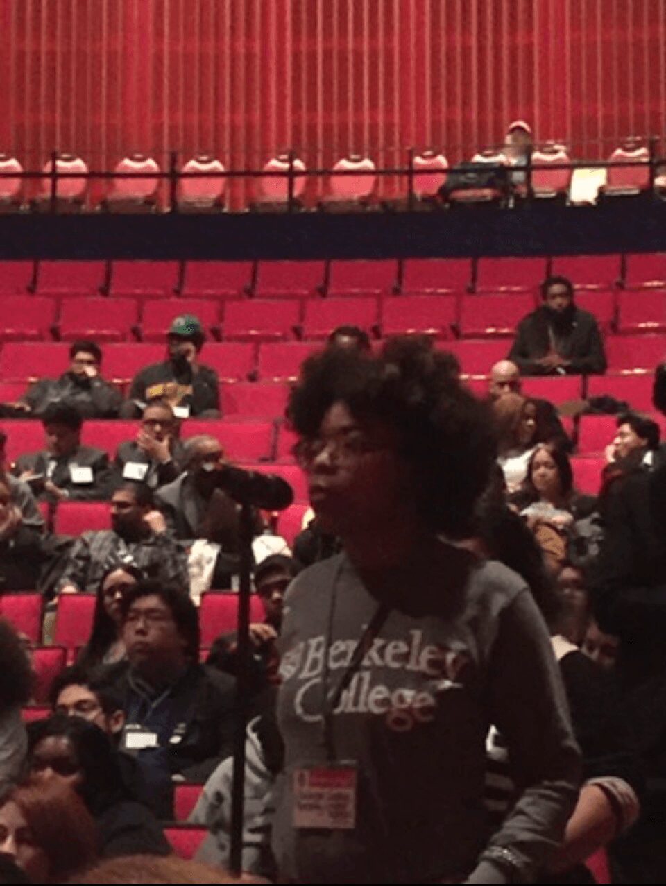 Person with curly hair and glasses standing in a large auditorium with red seats and audience members.