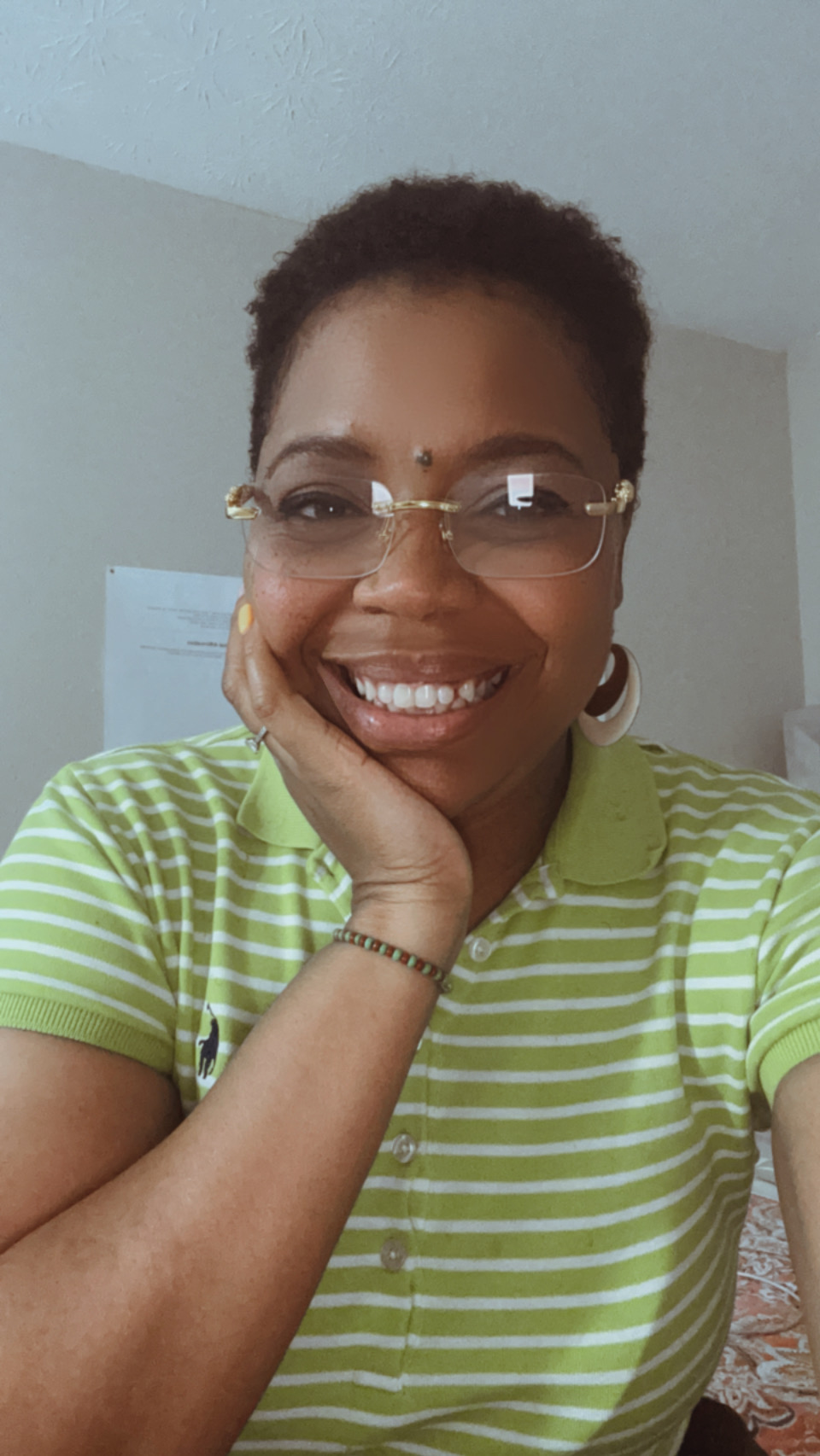 Woman with short curly hair, glasses, smiling, wearing a green and white striped shirt, resting her chin on her hand.
