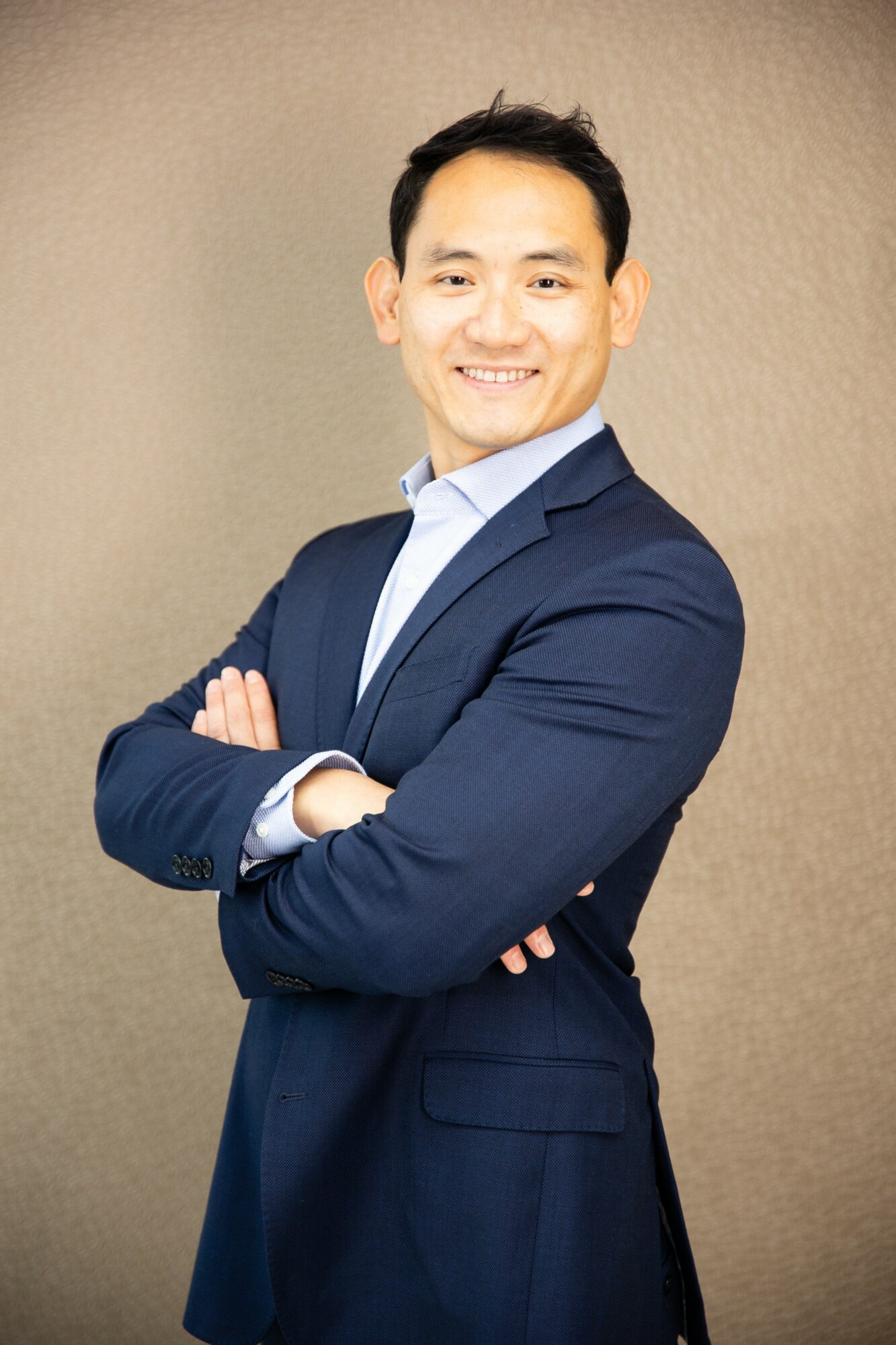 Man in a navy suit with arms crossed, smiling, against a plain background.