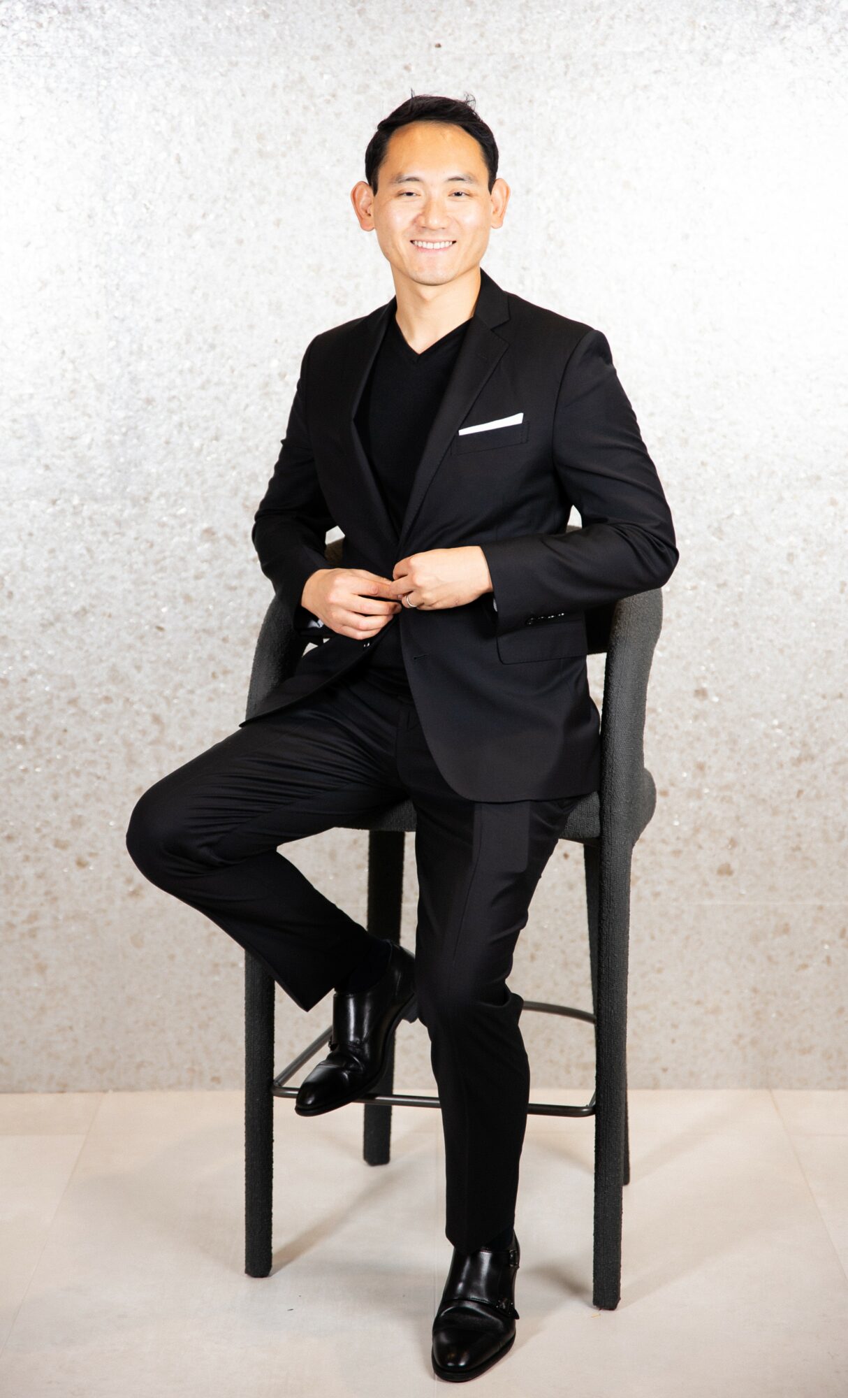 Young man in black suit sitting on a high chair, smiling, against a light textured background.