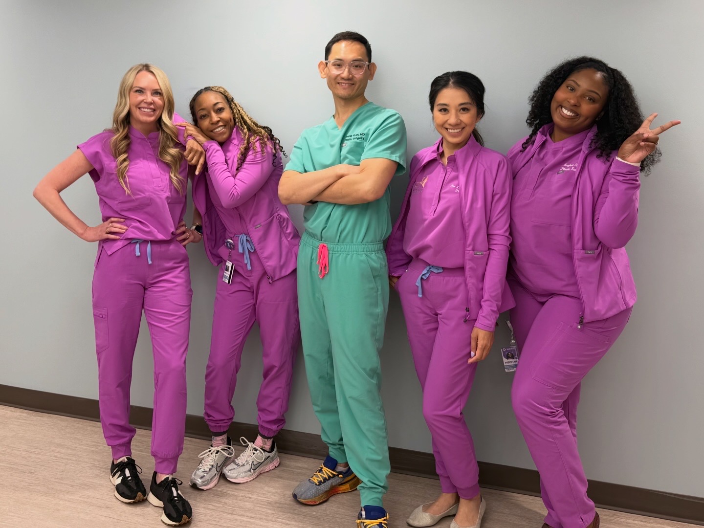 Group of five healthcare professionals in scrubs standing against a plain wall, smiling and posing casually.