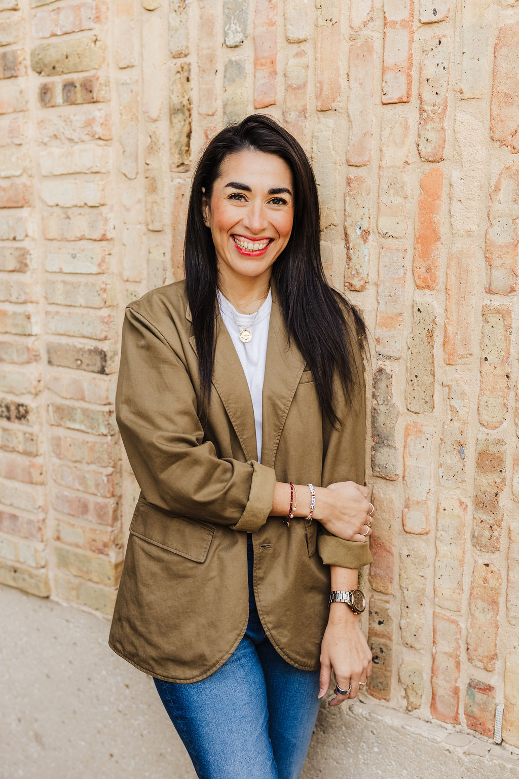 Woman with long dark hair smiling, wearing a brown blazer and blue jeans, standing against a brick wall.