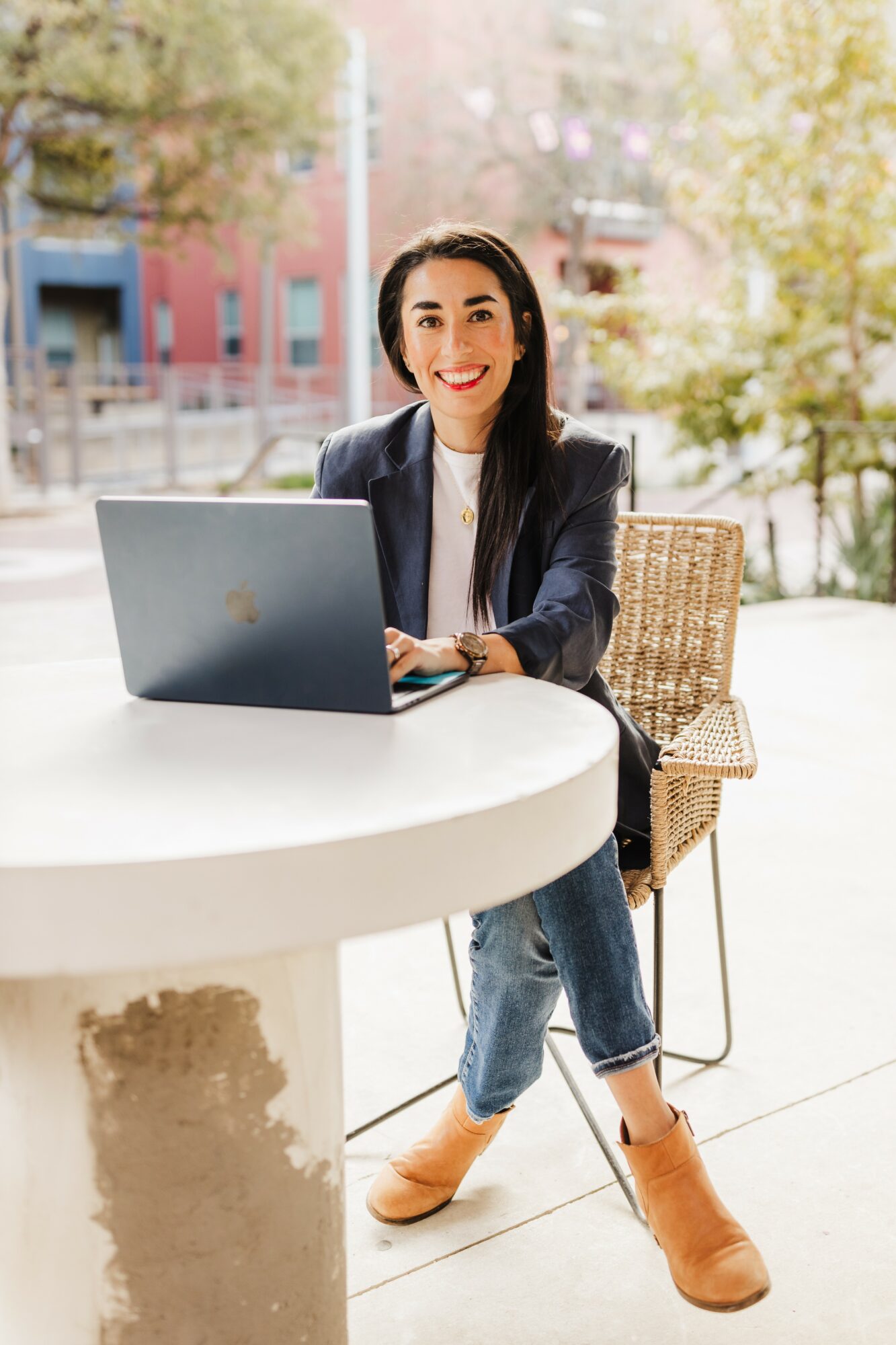 Woman sitting at a round table with a laptop outdoors, smiling, with trees and buildings in the background.