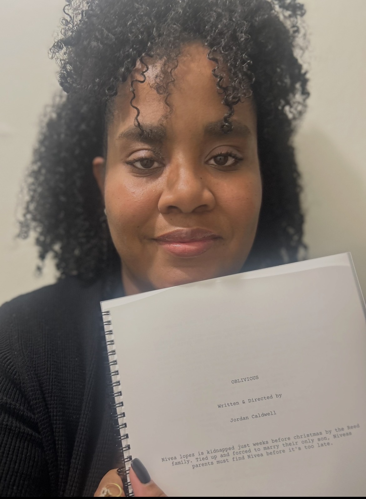 Woman with curly hair holding a blank notebook, looking at the camera, indoors.