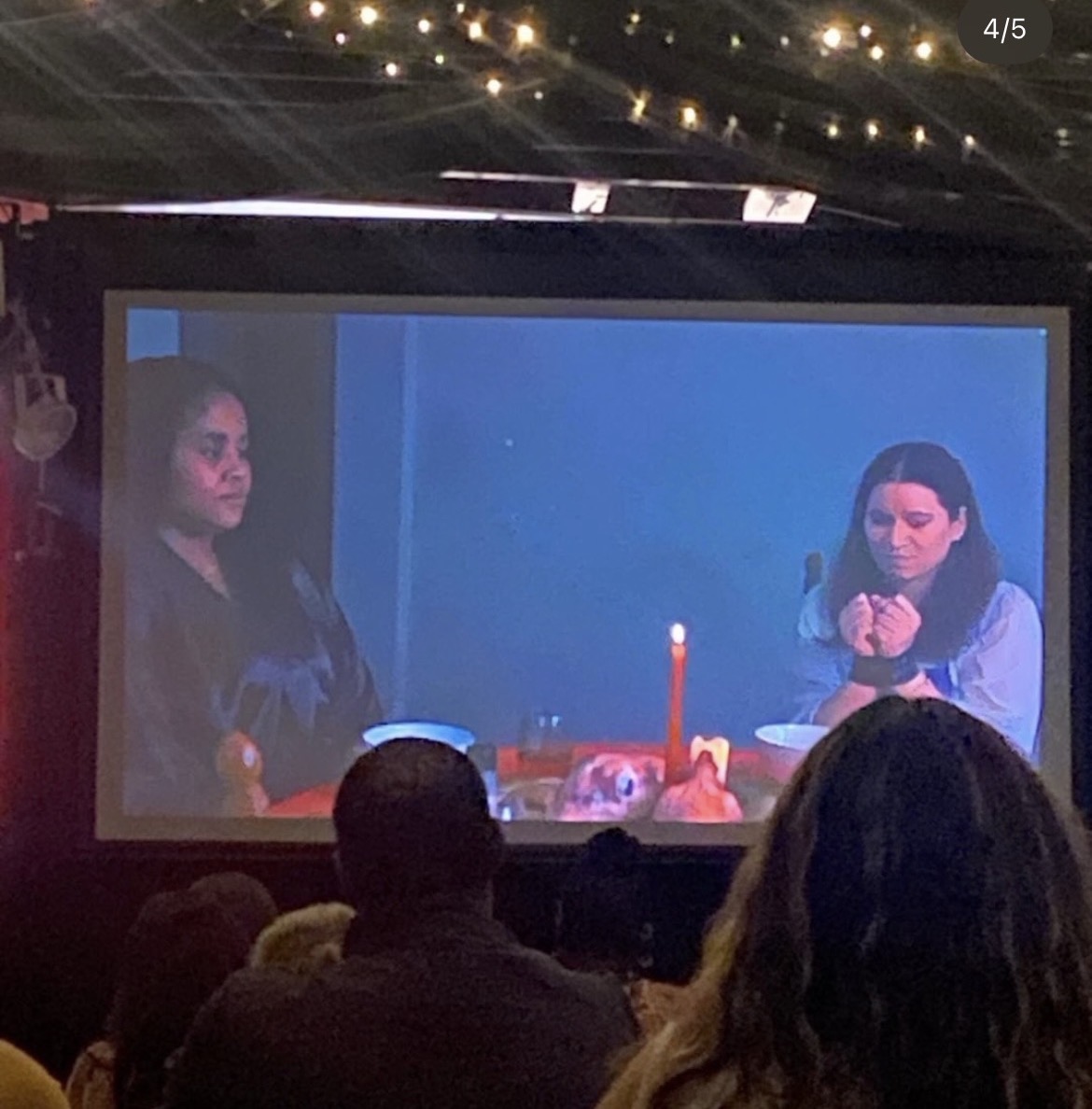 Two women sit at a table with a lit candle and food, watching a screen with a blue background, in a dark room.