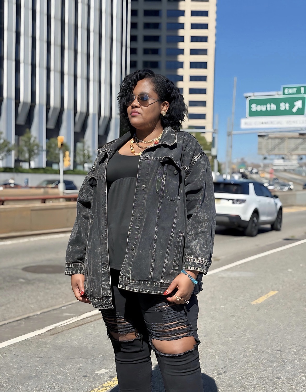 Woman standing on city street with tall buildings and cars, wearing sunglasses, black top, and ripped jeans.