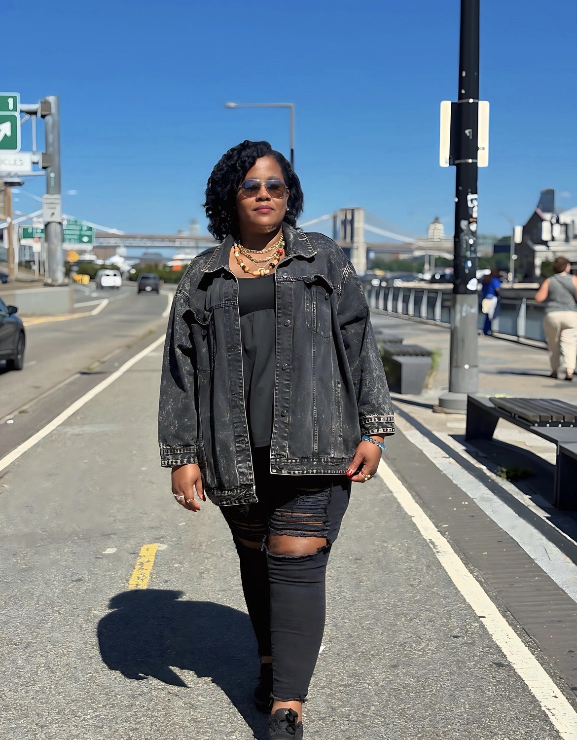 Woman walking on sidewalk in urban area, wearing black outfit and sunglasses, with bridge and cityscape in background.