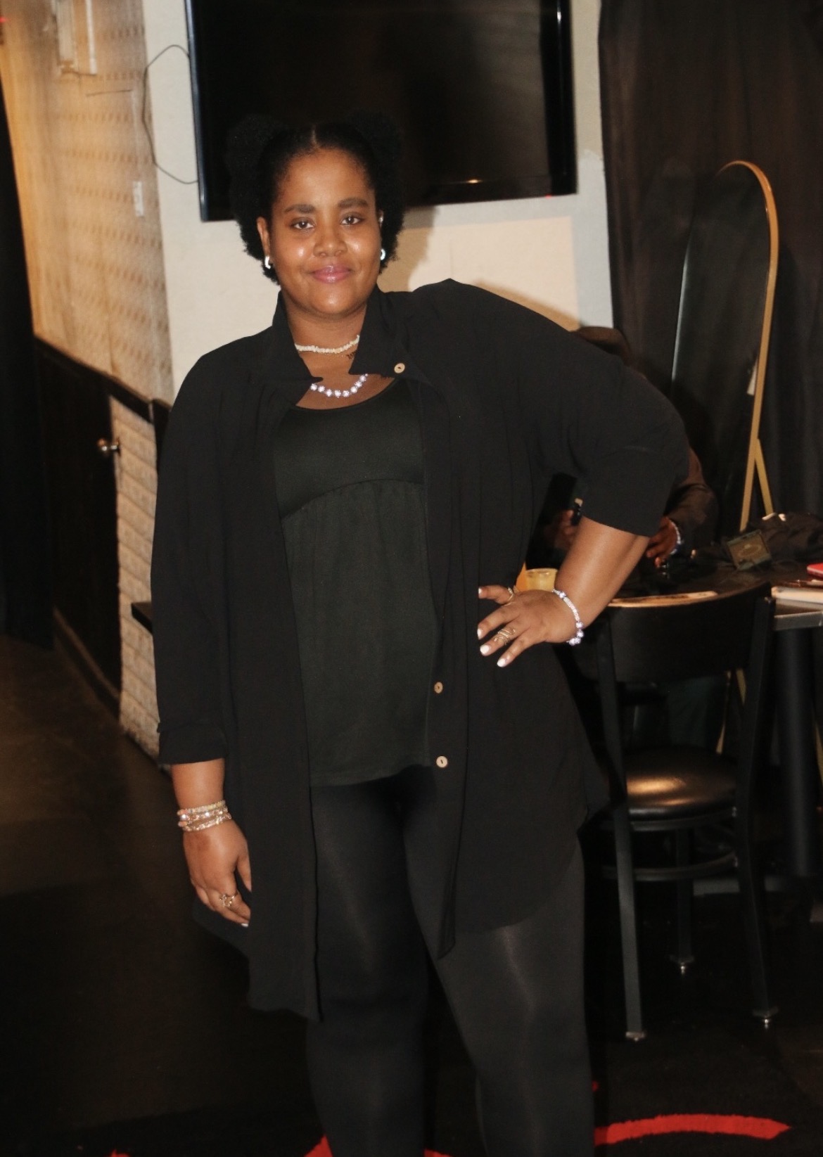 Woman standing indoors, wearing black outfit with jewelry, smiling, with a dark background and chairs nearby.