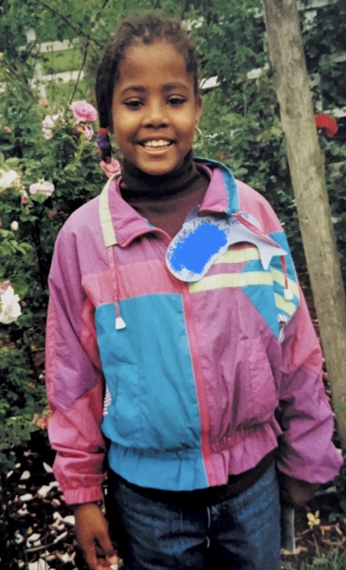 Young girl smiling outdoors with trees and flowers in background, wearing a colorful jacket and jeans.