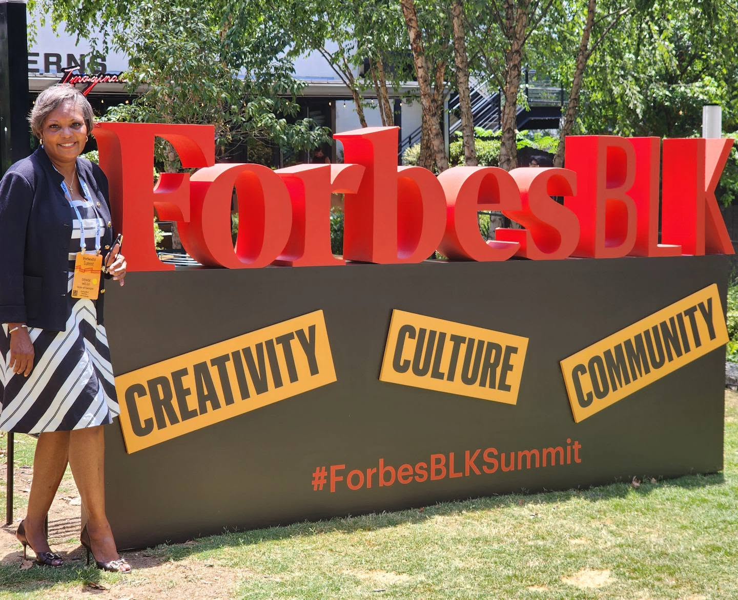 Woman standing next to large red ForbesBLK sign outdoors with trees and grass, wearing a striped dress and glasses.