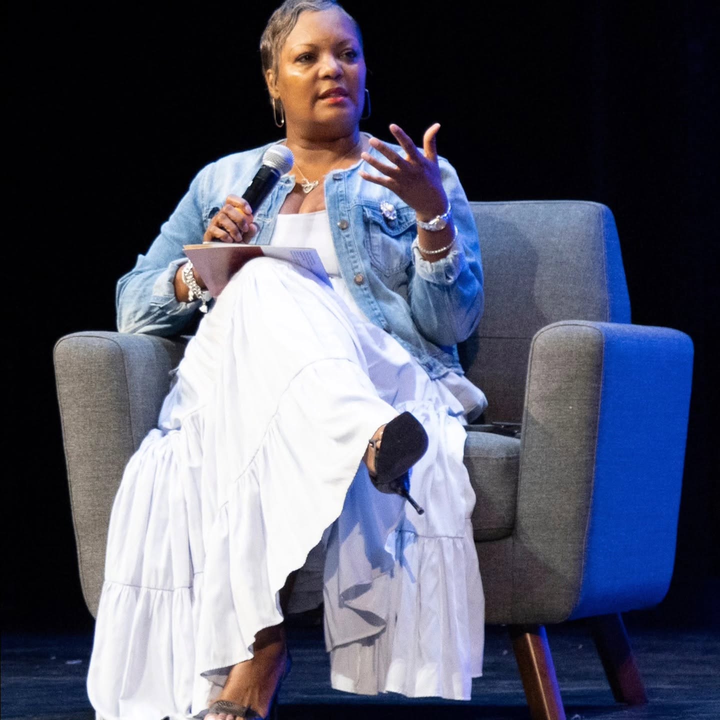 Woman sitting in a gray armchair, holding a microphone and gesturing with her hand, on a dark stage.
