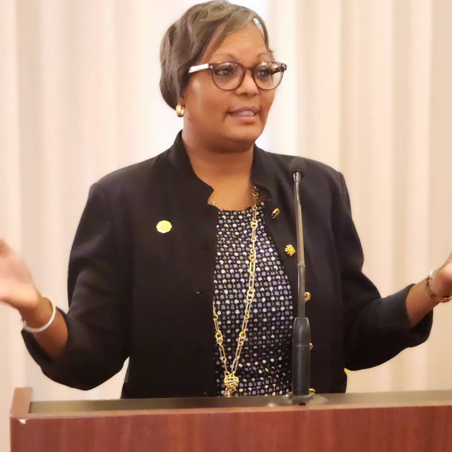Woman speaking at a podium with curtains in the background, wearing glasses, a black jacket, and a patterned top, gesturing with hands.