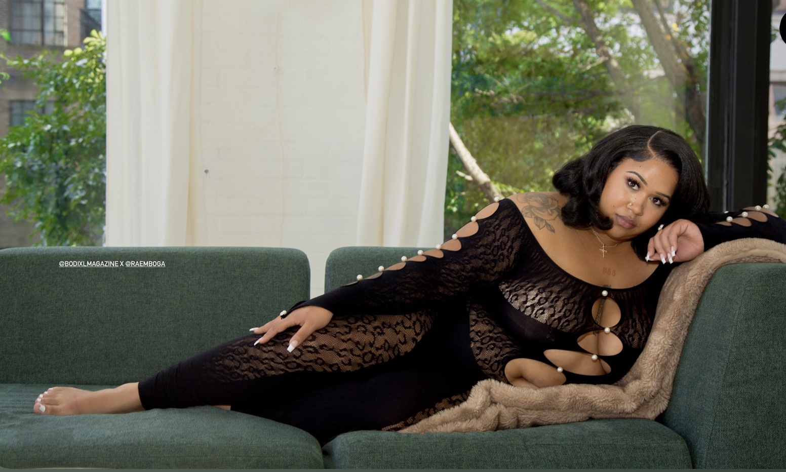 Woman in black lace dress lying on a green sofa near a window with trees outside.
