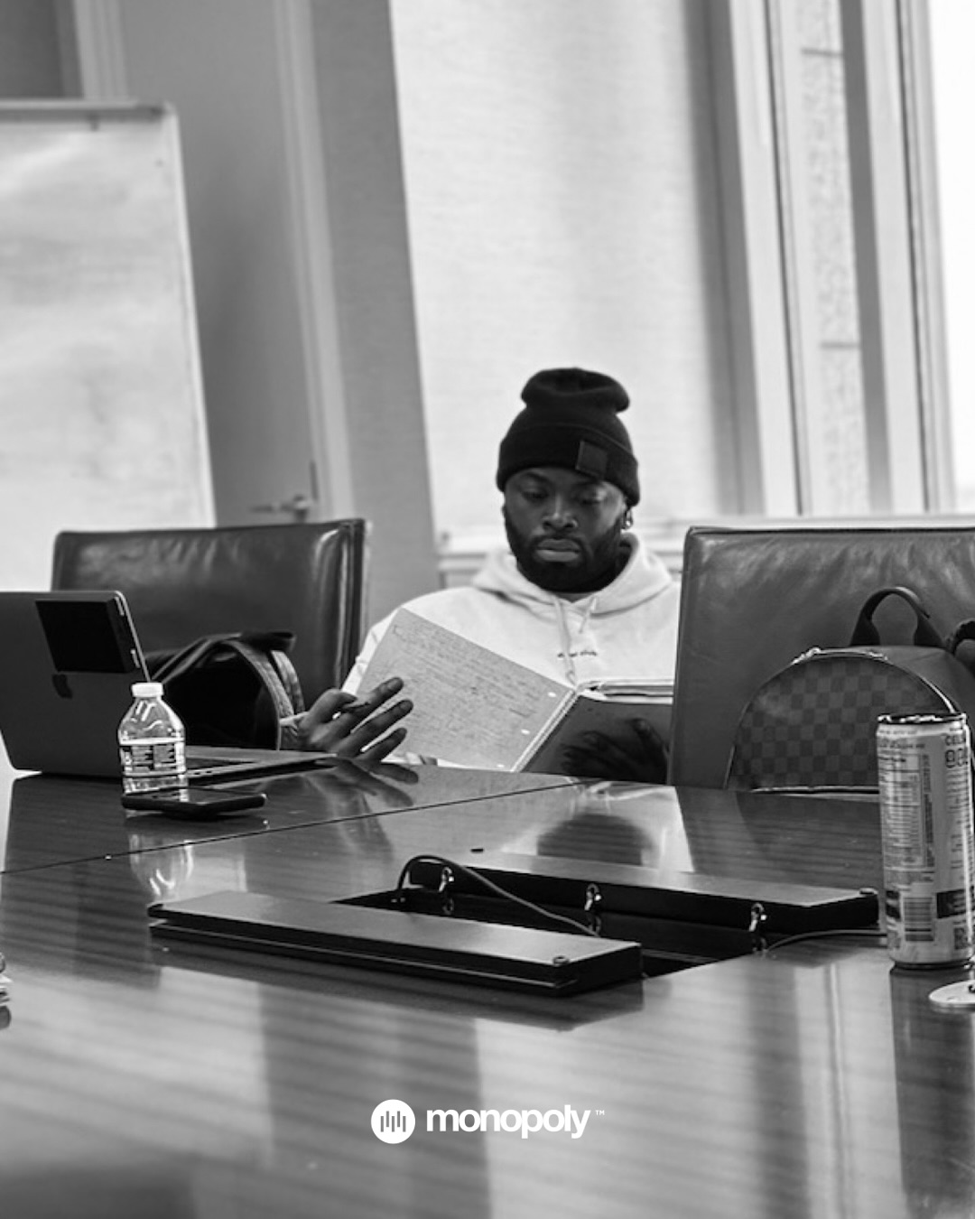 Man reading a book at a conference table with laptops, water bottles, and a window in the background.