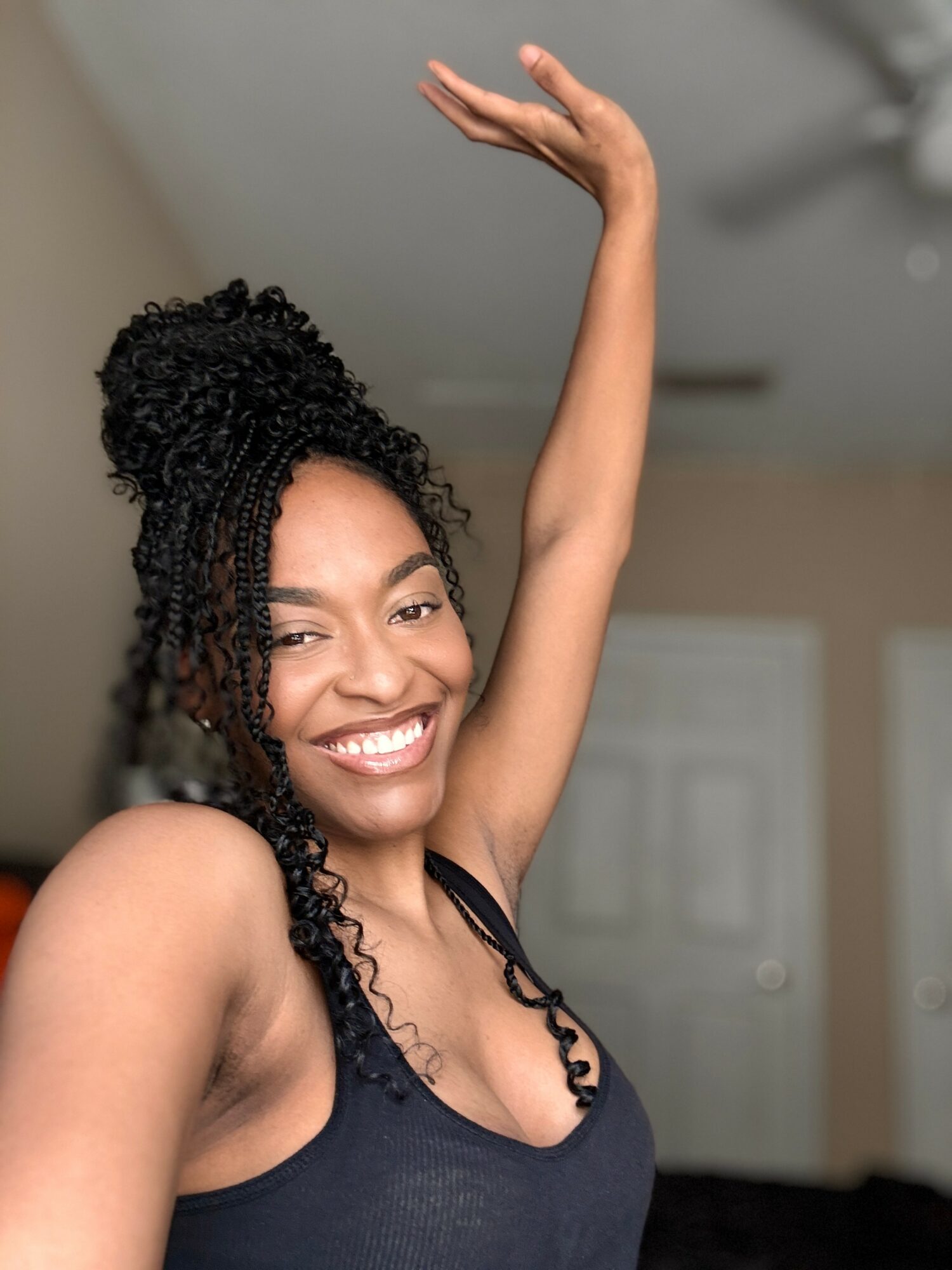 Smiling woman with curly hair raising her arm indoors, background with door and ceiling fan, casual attire.
