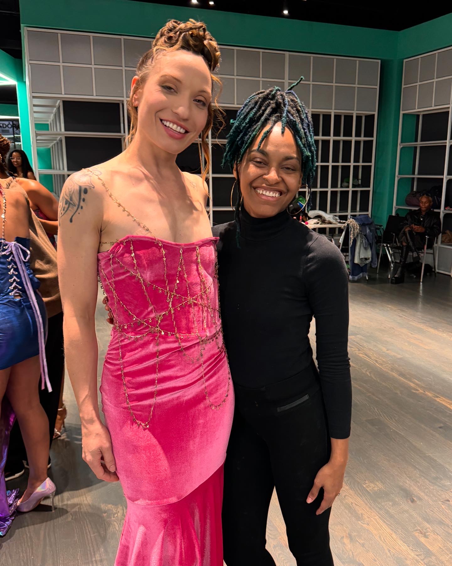 Two women smiling, one in a pink dress and the other in black, standing indoors with a group in the background.