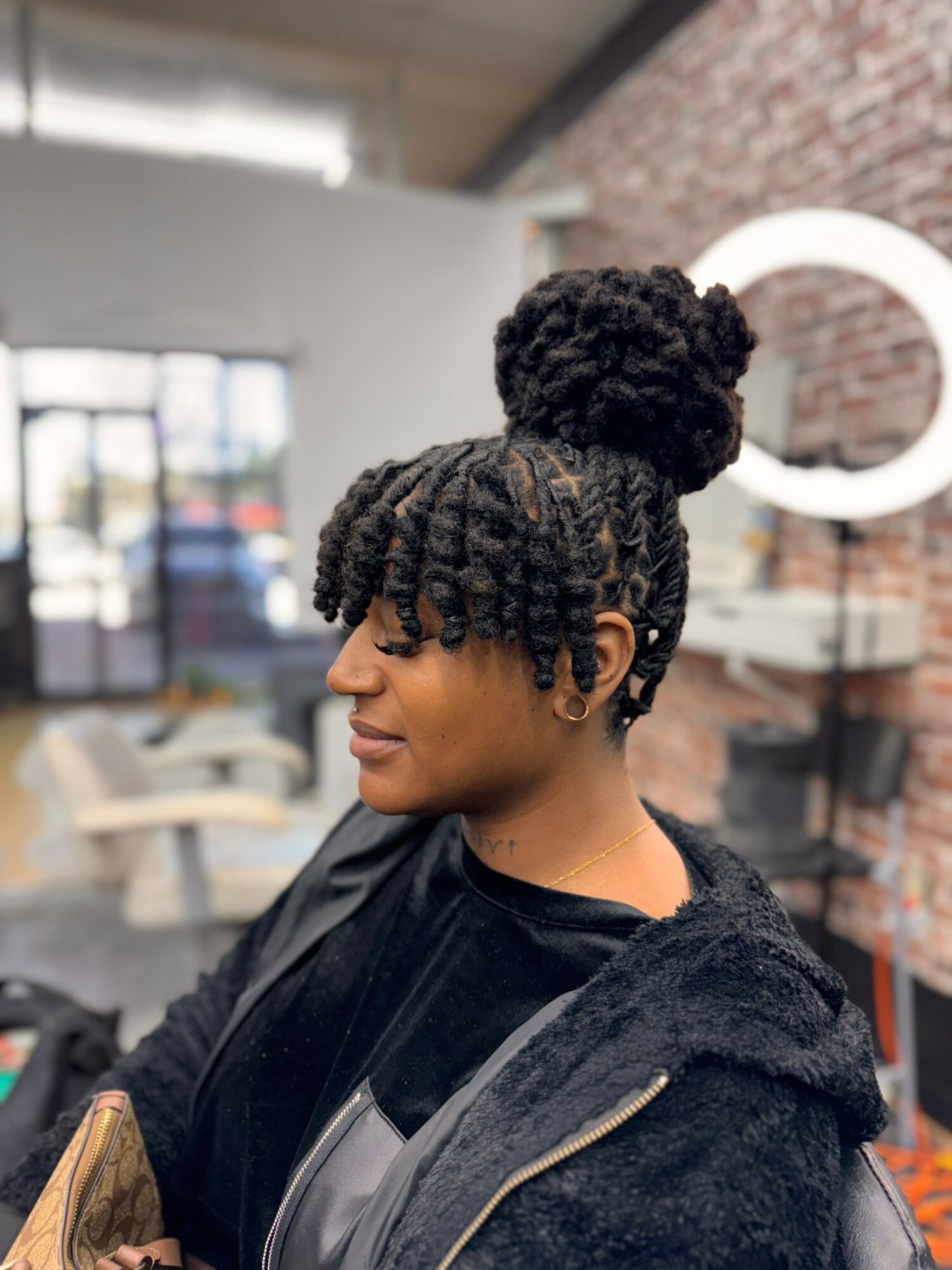 Side profile of a woman with styled natural hair in an indoor setting, brick wall and window in background.