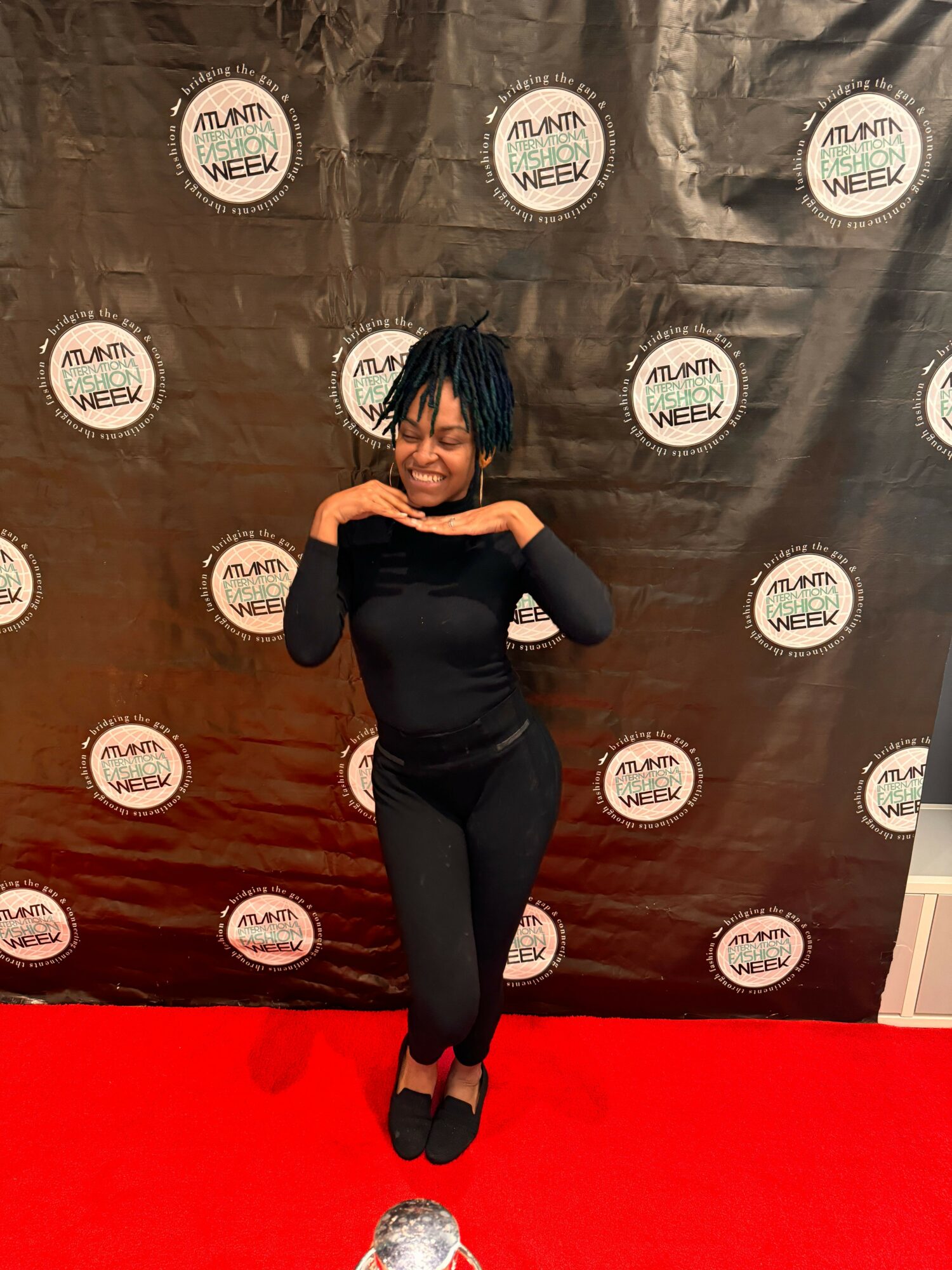 Woman in black dress and heels standing on red carpet with black backdrop behind her, smiling and posing with hands on hips.