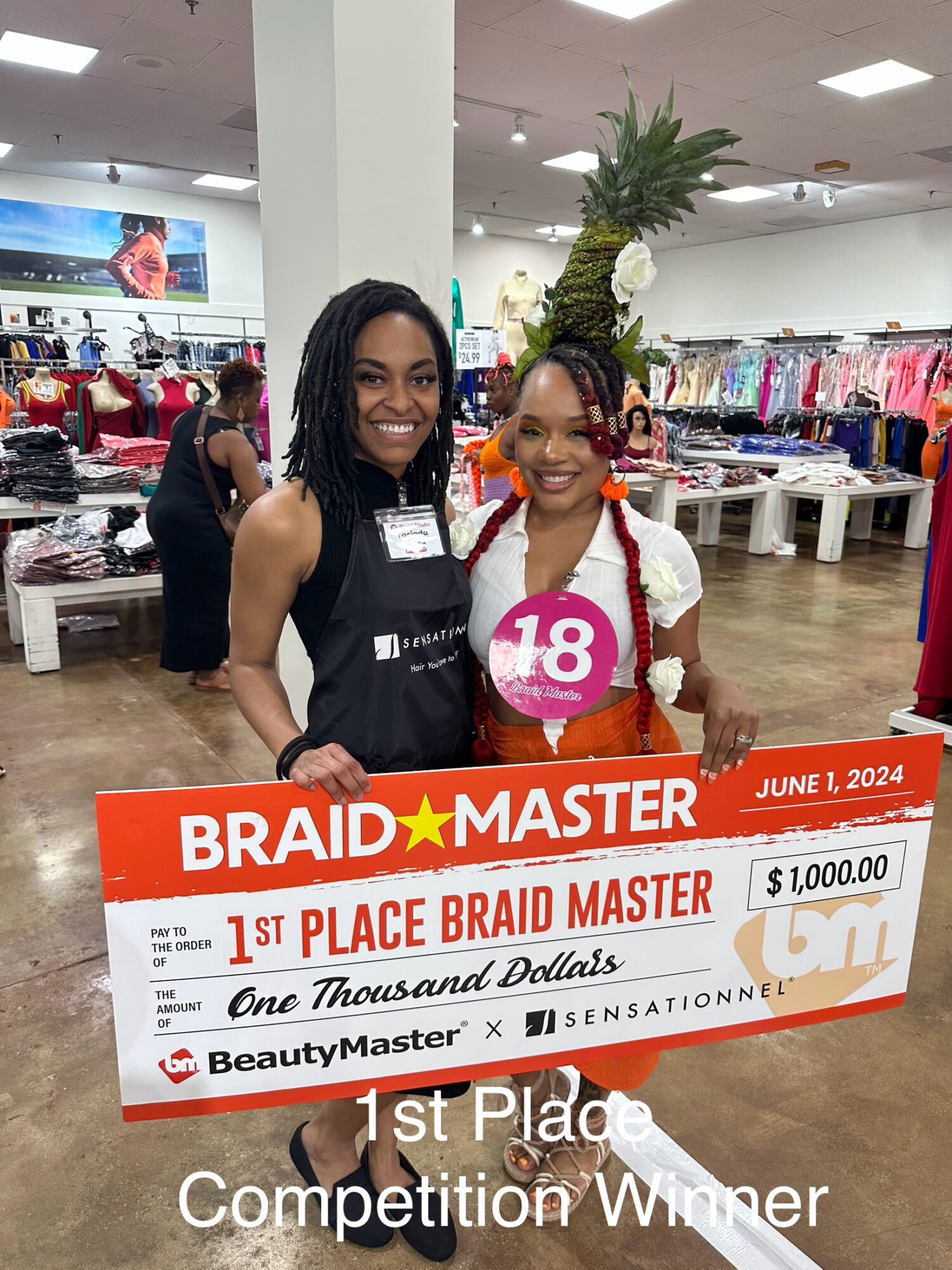 Two women smiling in a store, holding a large check for first place in a braid master competition, June 1, 2024.