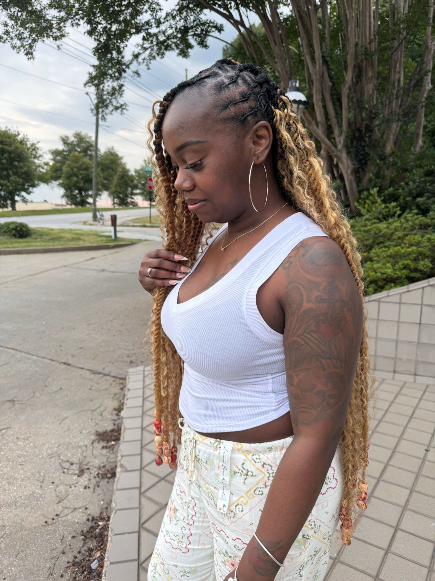 Woman with long braided hair, wearing a white tank top and patterned pants, standing outdoors near a tiled wall.