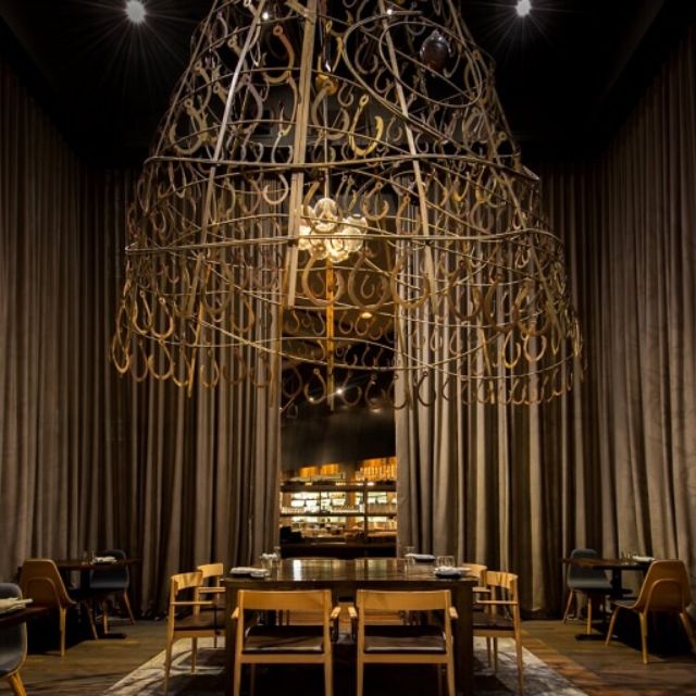 Large ornate chandelier hangs above a round table with chairs in a dimly lit room with curtains.