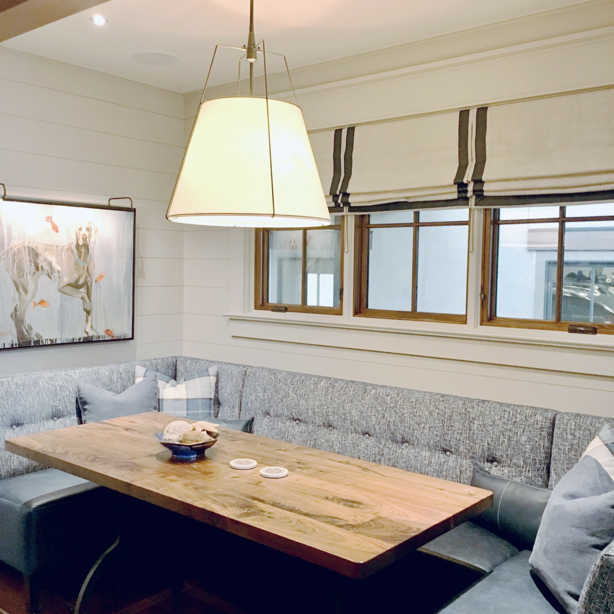 Corner of a room with a wooden table, gray upholstered seating, a window with blinds, and a hanging lamp.